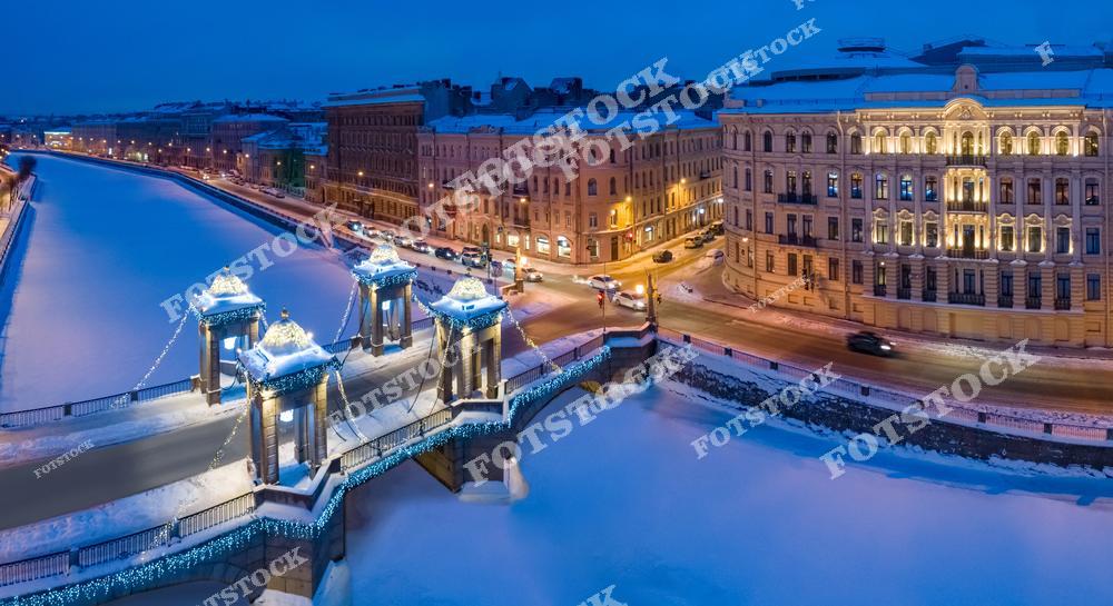 На фотографии изображён мост Ломоносова — один из самых красивых и узнаваемых мостов Санкт-Петербурга через реку Фонтанку. Построенный в XVIII веке, он сохранил подвижные пролёты и гранитные башни с цепями. В зимнее время мост украшен праздничной иллюминацией, придающей ему особое очарование. Замёрзшая Фонтанка и заснеженные улицы вокруг создают атмосферу тишины и уюта. Свет от фонарей и витрин отражается в снегу, усиливая ощущение праздника.